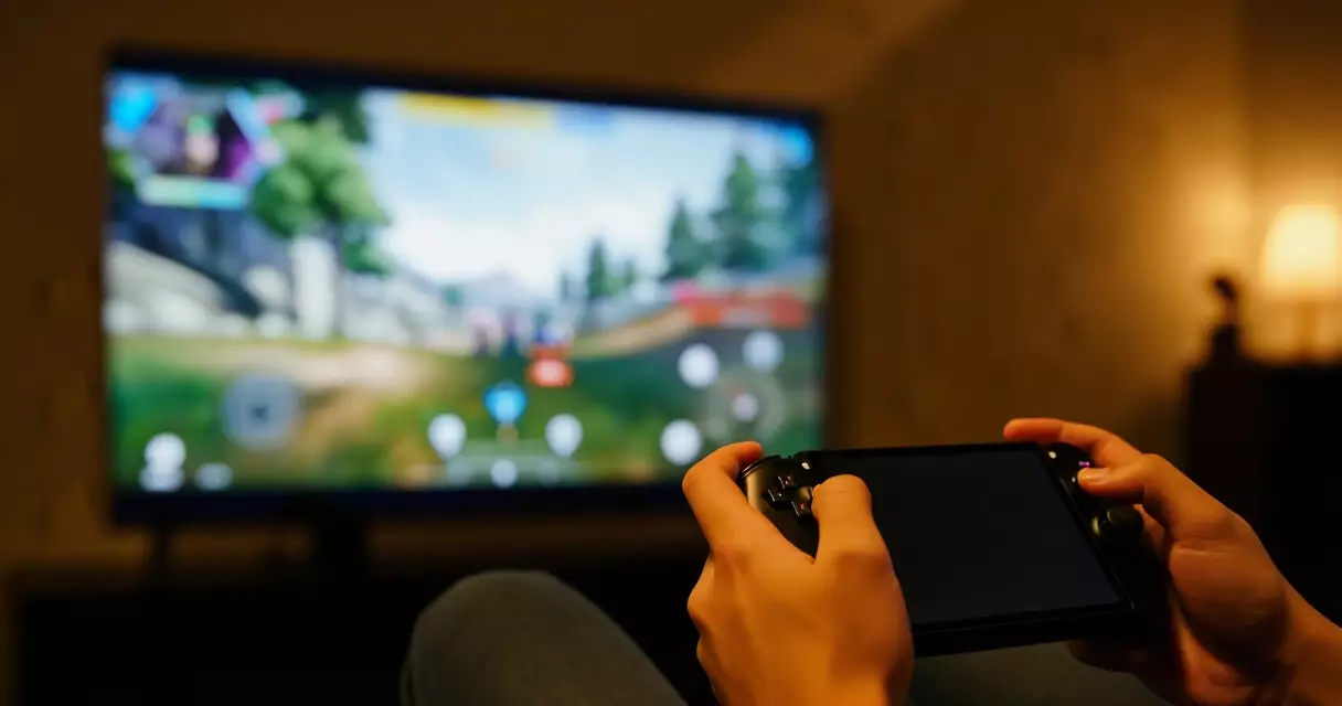 Gamer holding a handheld device with a console game paused on the TV behind
