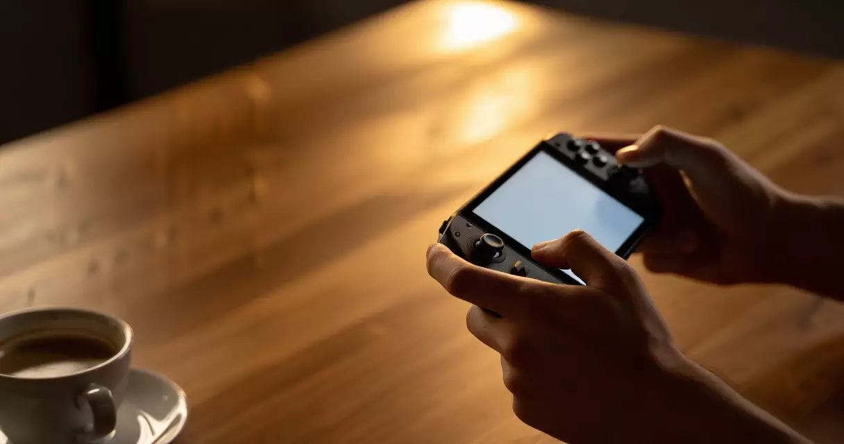 Hands holding a handheld gaming PC with dual thumbsticks and trackpads on a wooden desk